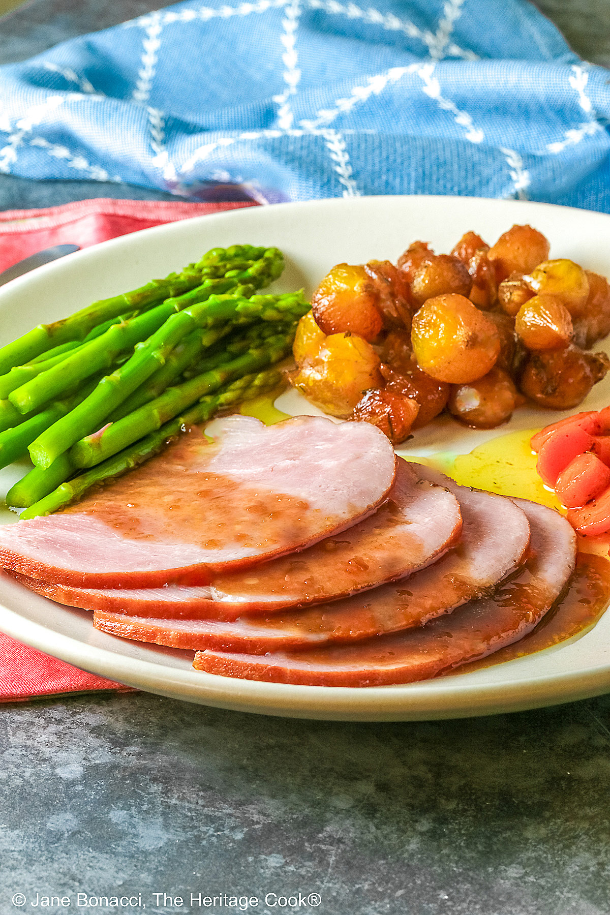 A pile of sliced Holiday Ham for Two topped with a maple-bourbon glaze on top, with spears of steamed asparagus, glazed pearl onions, and maple-dill carrots arranged around the ham slices on a white plate, sitting on a blue cloth with a pink napkin alongside © 2026 Jane Bonacci, The Heritage Cook.