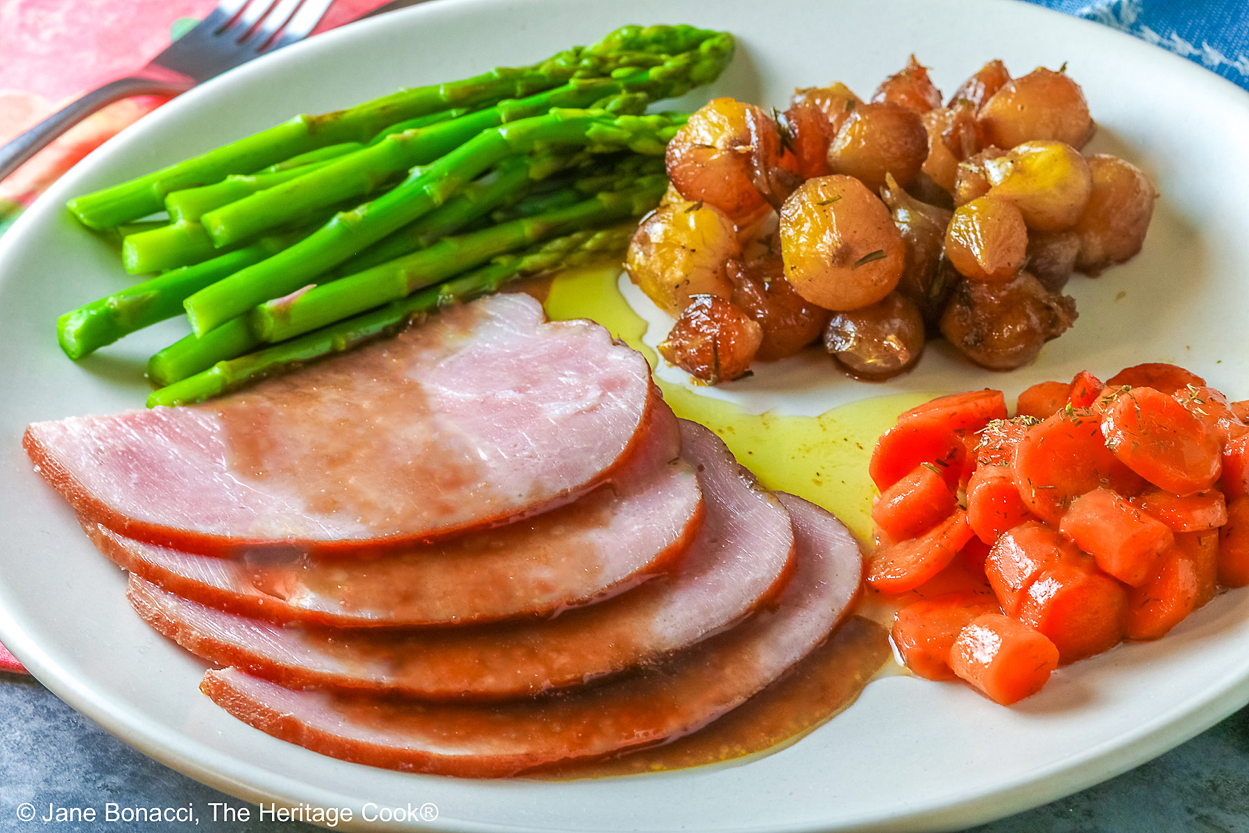 A pile of sliced Holiday Ham for Two topped with a maple-bourbon glaze on top, with spears of steamed asparagus, glazed pearl onions, and maple-dill carrots arranged around the ham slices on a white plate, sitting on a blue cloth with a pink napkin alongside © 2026 Jane Bonacci, The Heritage Cook.