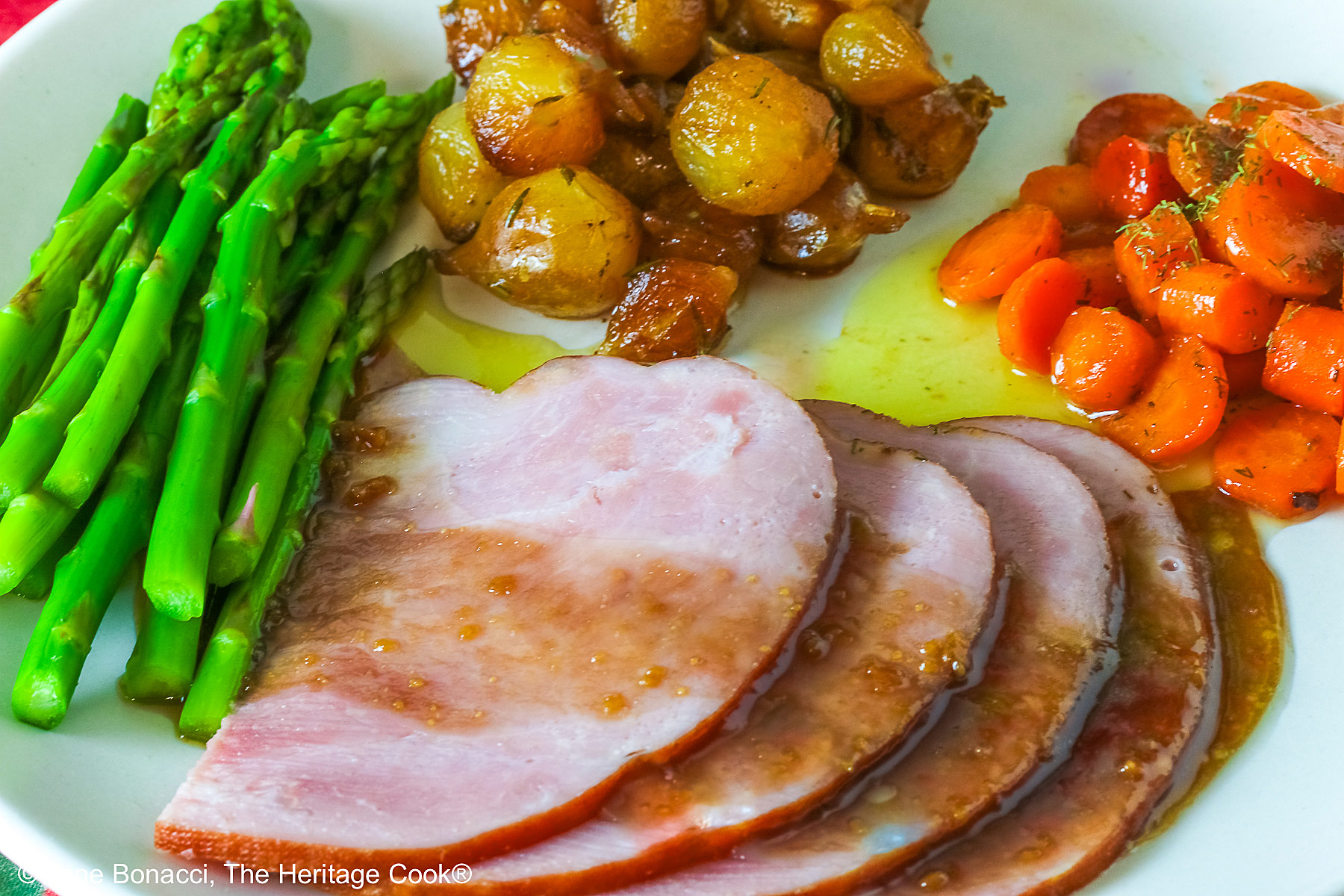A pile of sliced Holiday Ham for Two topped with a maple-bourbon glaze on top, with spears of steamed asparagus, glazed pearl onions, and maple-dill carrots arranged around the ham slices on a white plate, sitting on a blue cloth with a pink napkin alongside © 2026 Jane Bonacci, The Heritage Cook.