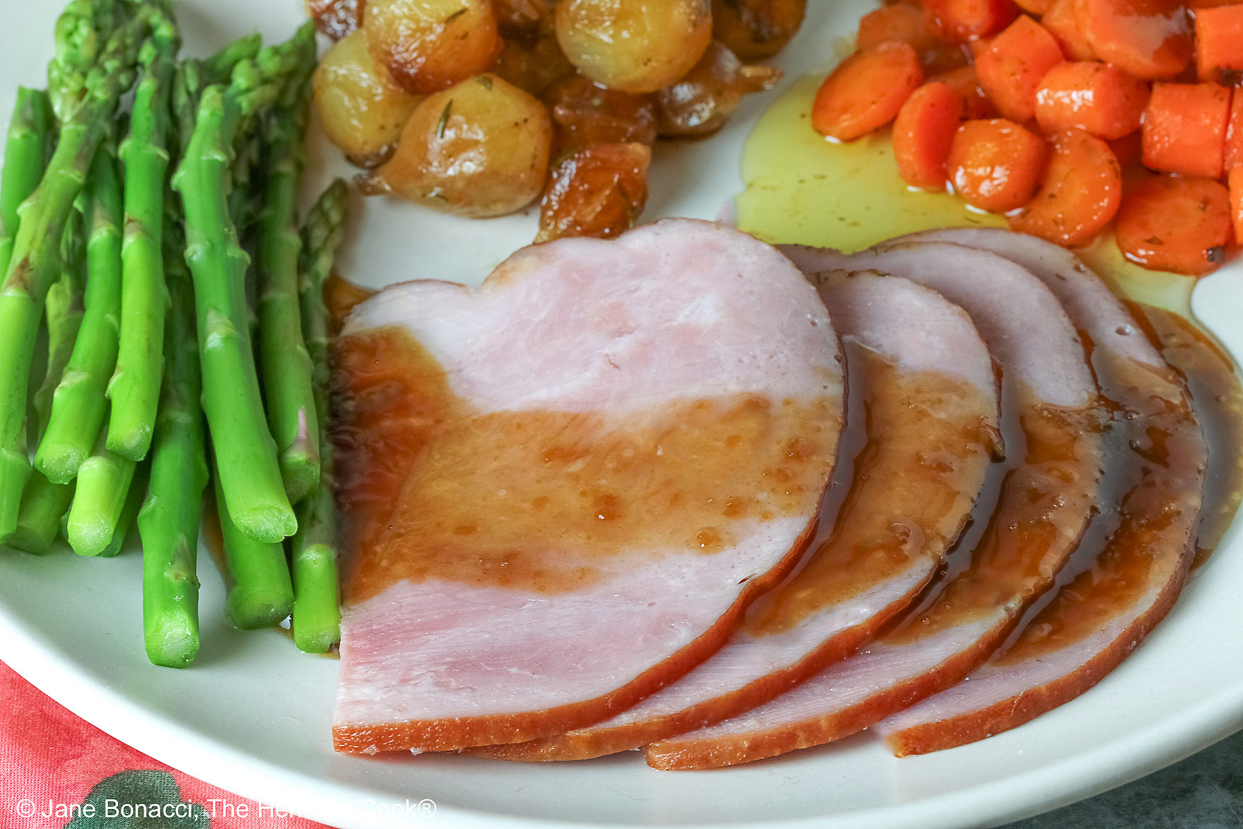 A pile of sliced Holiday Ham for Two topped with a maple-bourbon glaze on top, with spears of steamed asparagus, glazed pearl onions, and maple-dill carrots arranged around the ham slices on a white plate, sitting on a blue cloth with a pink napkin alongside © 2026 Jane Bonacci, The Heritage Cook.