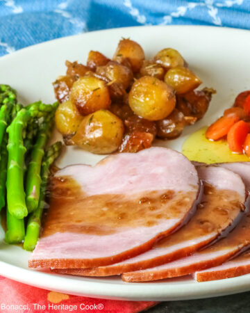 A pile of sliced Holiday Ham for Two topped with a maple-bourbon glaze on top, with spears of steamed asparagus, glazed pearl onions, and maple-dill carrots arranged around the ham slices on a white plate, sitting on a blue cloth with a pink napkin alongside © 2026 Jane Bonacci, The Heritage Cook.