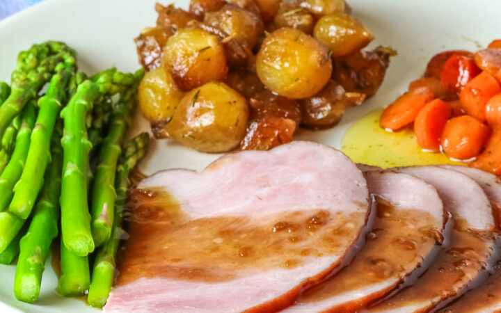 A pile of sliced Holiday Ham for Two topped with a maple-bourbon glaze on top, with spears of steamed asparagus, glazed pearl onions, and maple-dill carrots arranged around the ham slices on a white plate, sitting on a blue cloth with a pink napkin alongside © 2026 Jane Bonacci, The Heritage Cook.