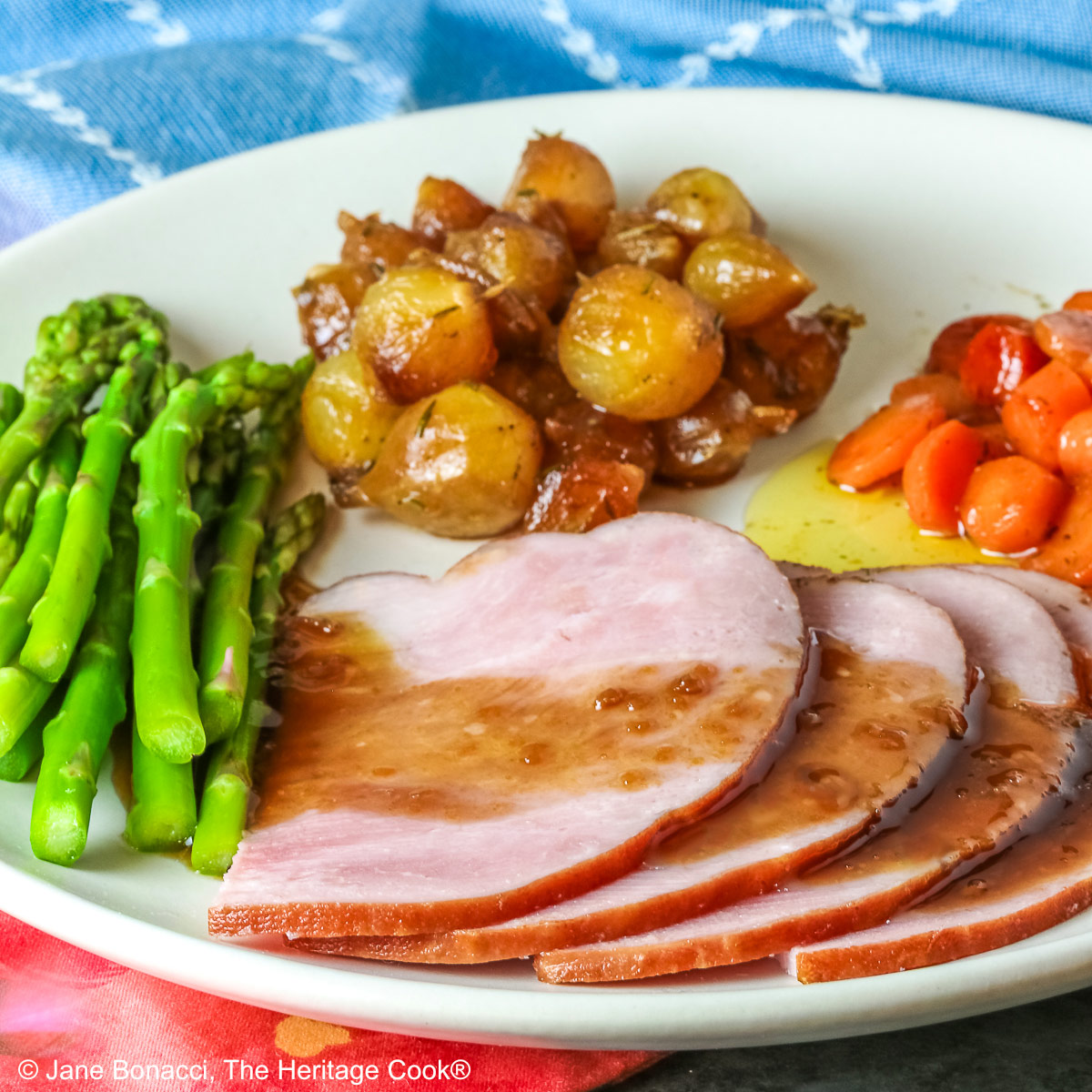 A pile of sliced Holiday Ham for Two topped with a maple-bourbon glaze on top, with spears of steamed asparagus, glazed pearl onions, and maple-dill carrots arranged around the ham slices on a white plate, sitting on a blue cloth with a pink napkin alongside © 2026 Jane Bonacci, The Heritage Cook.