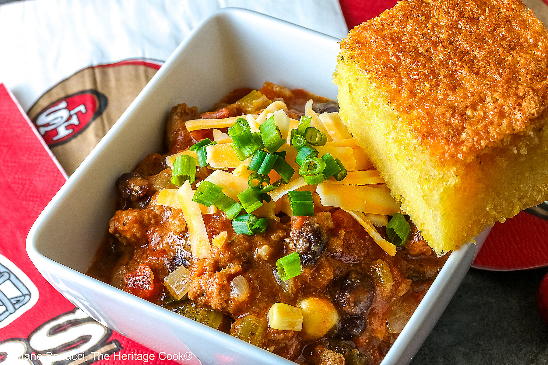 A square of Honey-Glazed Cornbread perched on the corner of a white square bowl surrounded with 49er football napkins and red and gold chocolate candies. The bowl is filled with Pork and Chorizo chili, topped with shredded cheese and chopped green onions on a gray background © 2026 Jane Bonacci, The Heritage Cook.