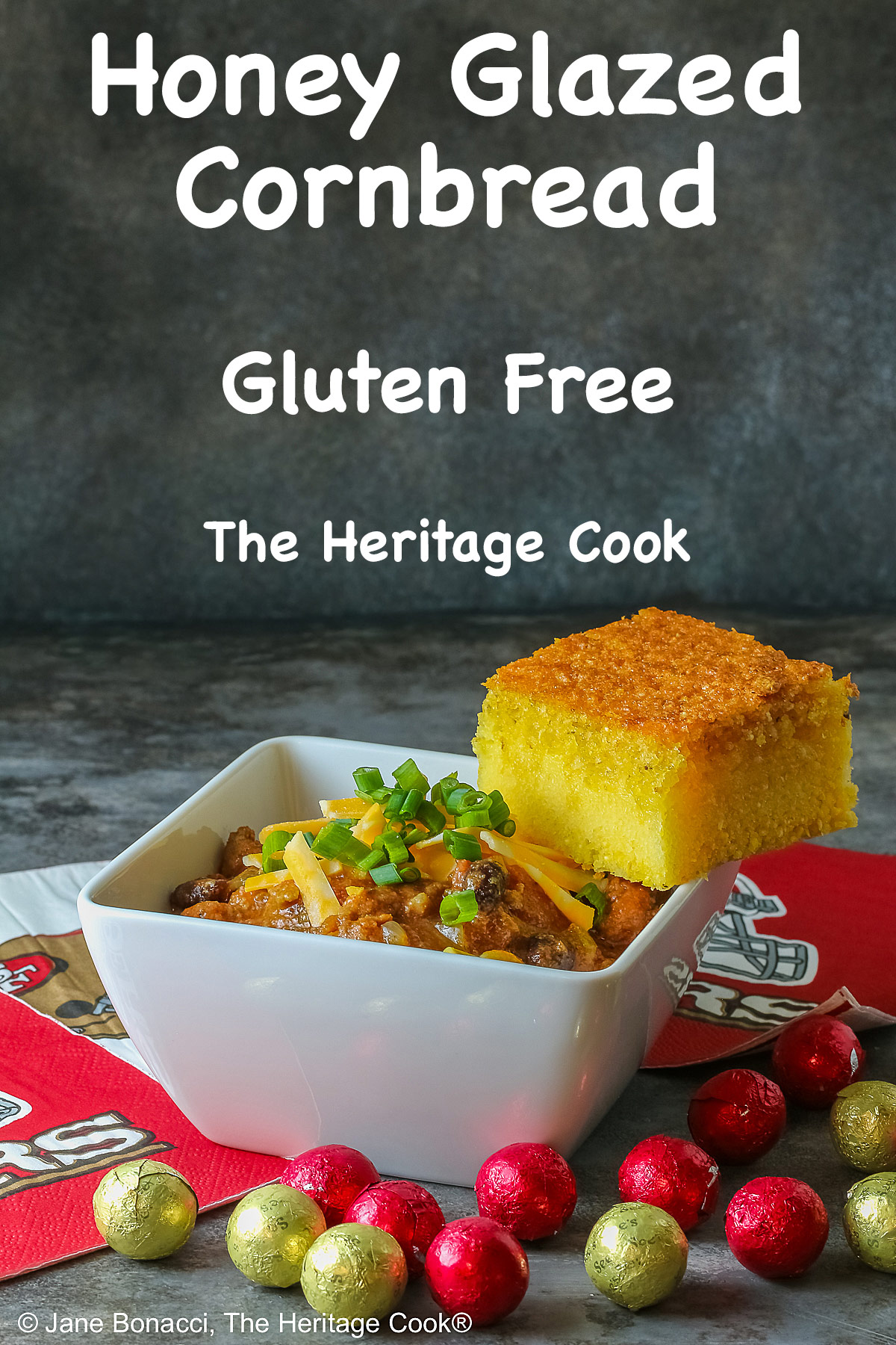 A square of Honey-Glazed Cornbread perched on the corner of a white square bowl surrounded with 49er football napkins and red and gold chocolate candies. The bowl is filled with Pork and Chorizo chili, topped with shredded cheese and chopped green onions on a gray background © 2026 Jane Bonacci, The Heritage Cook.