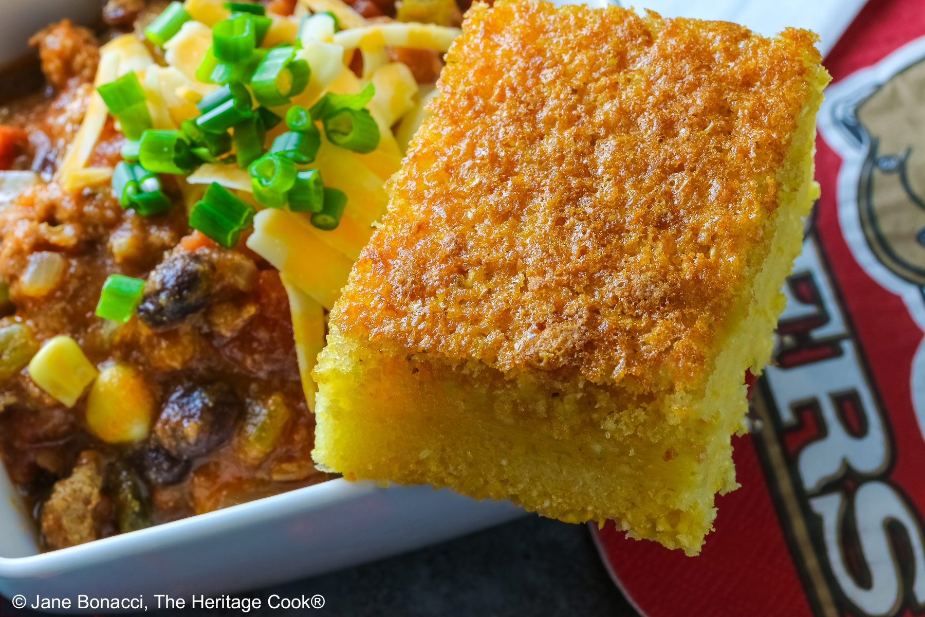 A square of Honey-Glazed Cornbread perched on the corner of a white square bowl surrounded with 49er football napkins and red and gold chocolate candies. The bowl is filled with Pork and Chorizo chili, topped with shredded cheese and chopped green onions on a gray background © 2026 Jane Bonacci, The Heritage Cook.