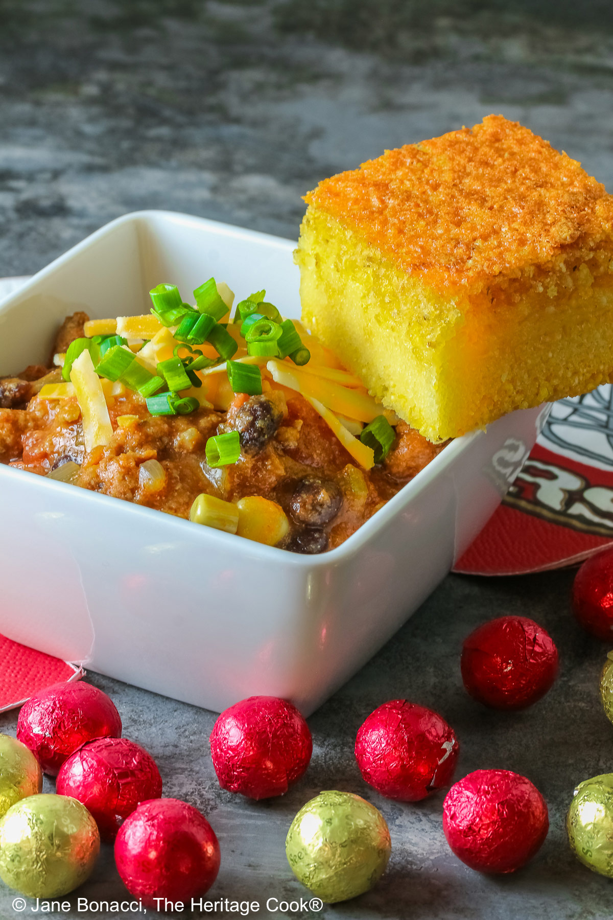 A square of Honey-Glazed Cornbread perched on the corner of a white square bowl surrounded with 49er football napkins and red and gold chocolate candies. The bowl is filled with Pork and Chorizo chili, topped with shredded cheese and chopped green onions on a gray background © 2026 Jane Bonacci, The Heritage Cook.
