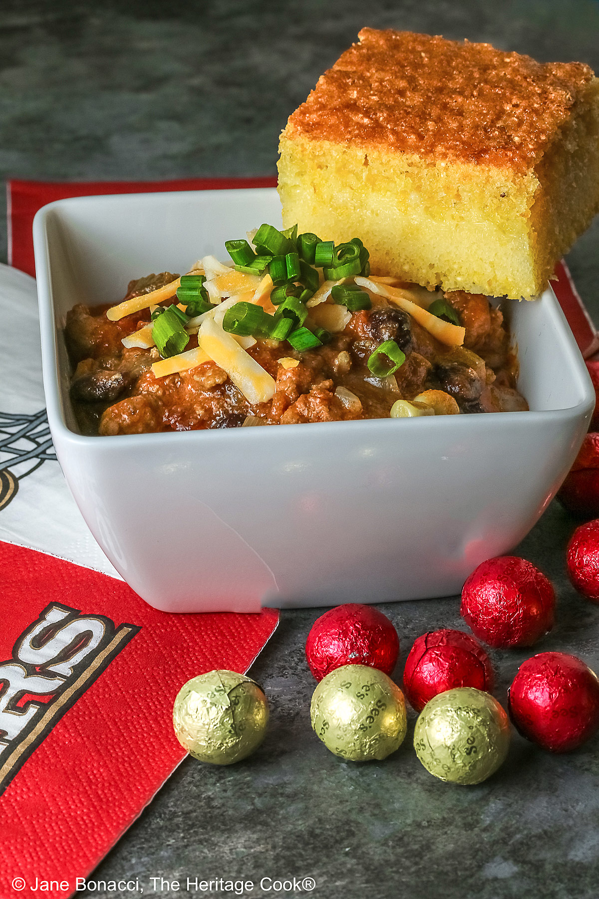 A square of Honey-Glazed Cornbread perched on the corner of a white square bowl surrounded with 49er football napkins and red and gold chocolate candies. The bowl is filled with Pork and Chorizo chili, topped with shredded cheese and chopped green onions on a gray background © 2026 Jane Bonacci, The Heritage Cook.