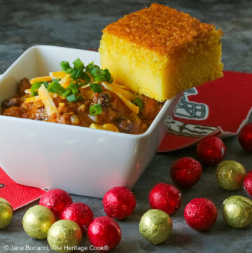 A square of Honey-Glazed Cornbread perched on the corner of a white square bowl surrounded with 49er football napkins and red and gold chocolate candies. The bowl is filled with Pork and Chorizo chili, topped with shredded cheese and chopped green onions on a gray background © 2026 Jane Bonacci, The Heritage Cook.