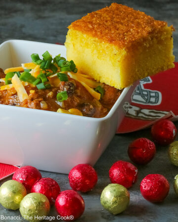 A square of Honey-Glazed Cornbread perched on the corner of a white square bowl surrounded with 49er football napkins and red and gold chocolate candies. The bowl is filled with Pork and Chorizo chili, topped with shredded cheese and chopped green onions on a gray background © 2026 Jane Bonacci, The Heritage Cook.