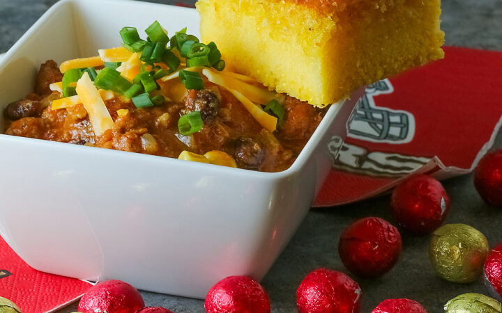 A square of Honey-Glazed Cornbread perched on the corner of a white square bowl surrounded with 49er football napkins and red and gold chocolate candies. The bowl is filled with Pork and Chorizo chili, topped with shredded cheese and chopped green onions on a gray background © 2026 Jane Bonacci, The Heritage Cook.
