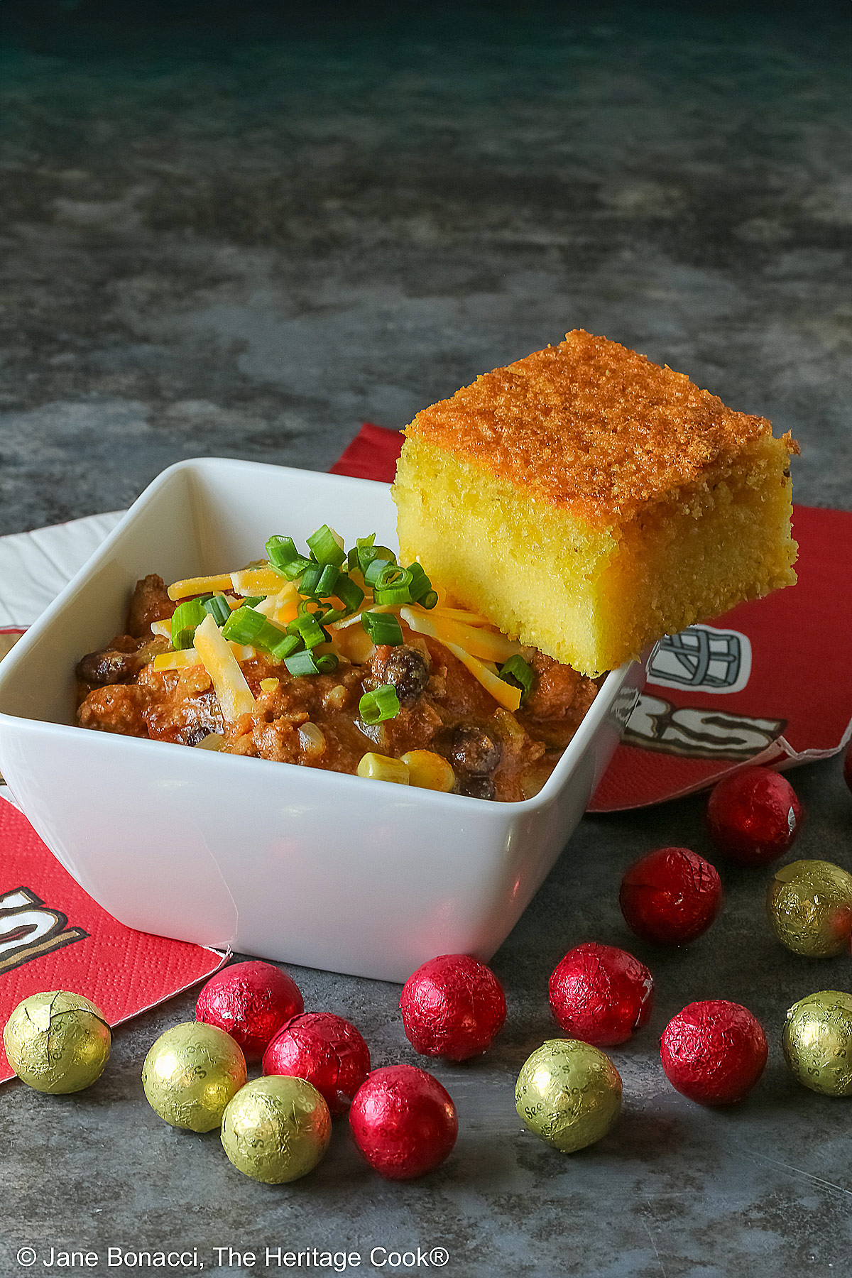 A square of Honey-Glazed Cornbread perched on the corner of a white square bowl surrounded with 49er football napkins and red and gold chocolate candies. The bowl is filled with Pork and Chorizo chili, topped with shredded cheese and chopped green onions on a gray background © 2026 Jane Bonacci, The Heritage Cook.