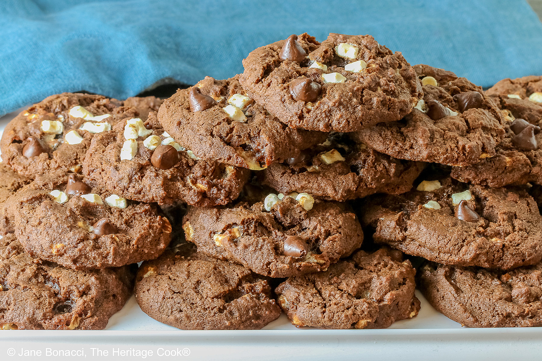 Hot Cocoa Cookies stacked in a wide pyramid shape, each cookie topped with extra micro mini marshmallow bits and chocolate chips, a big pile of dark brown chocolate cookies © 2026 Jane Bonacci, The Heritage Cook.