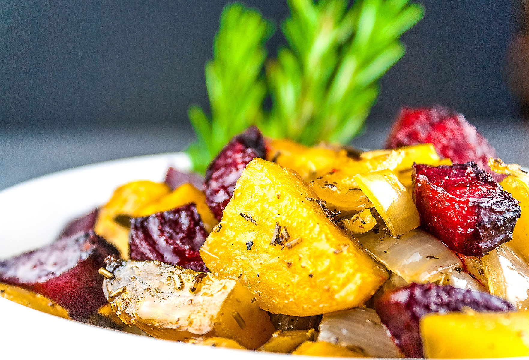 A mélange of roasted beets, potatoes, and peppers piled in a red au gratin baking dish or white baking dish with a sprig of rosemary in the back, placed on a white surface with a black or blue background © 2026 Jane Bonacci, The Heritage Cook.