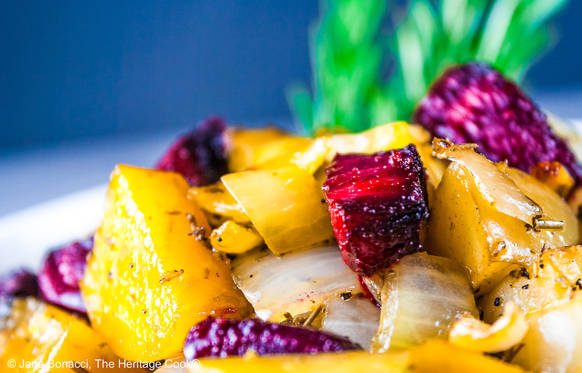 A mélange of roasted beets, potatoes, and peppers piled in a red au gratin baking dish or white baking dish with a sprig of rosemary in the back, placed on a white surface with a black or blue background © 2026 Jane Bonacci, The Heritage Cook.