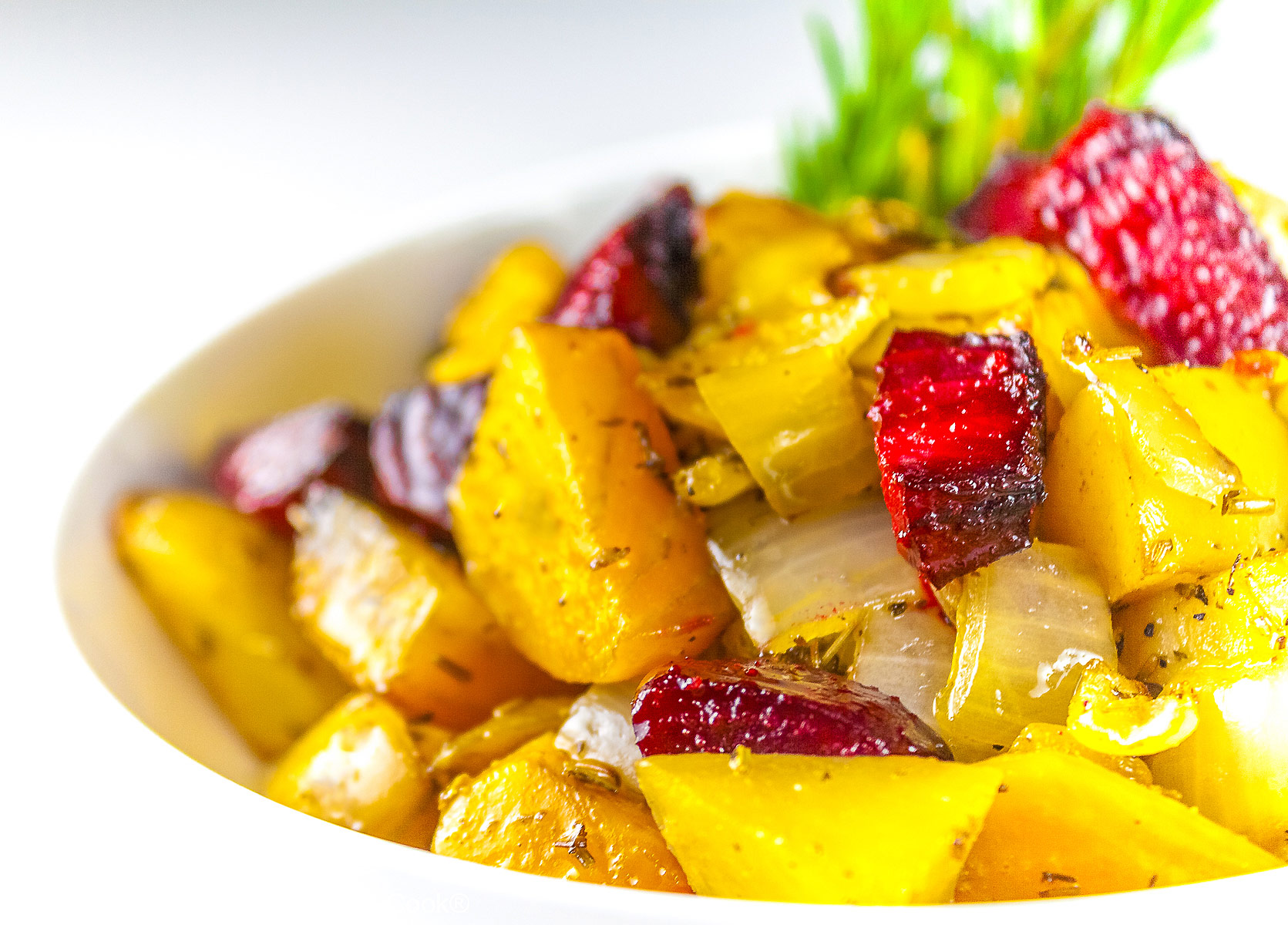 A mélange of roasted beets, potatoes, and peppers piled in a red au gratin baking dish or white baking dish with a sprig of rosemary in the back, placed on a white surface with a black or blue background © 2026 Jane Bonacci, The Heritage Cook.