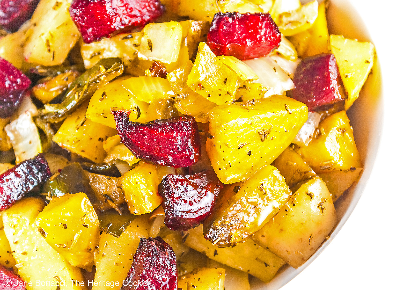 A mélange of roasted beets, potatoes, and peppers piled in a red au gratin baking dish or white baking dish with a sprig of rosemary in the back, placed on a white surface with a black or blue background © 2026 Jane Bonacci, The Heritage Cook.