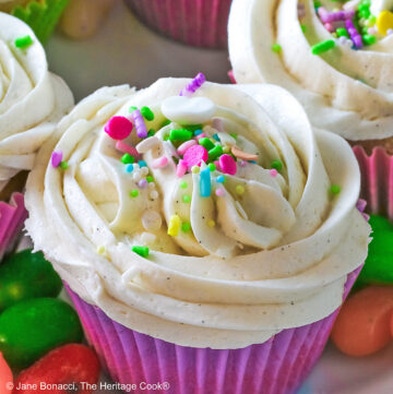 White Chocolate Cupcakes with piped white buttercream frosting, topped with Easter pastel sprinkles in festive pastel Spring colored paper liners, surrounded by brightly colored jelly beans and little fluffy chick and bunny on the side, all sitting on a deep pink cloth © 2026 Jane Bonacci, The Heritage Cook.