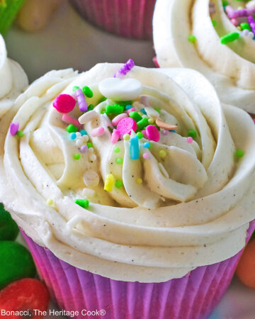 White Chocolate Cupcakes with piped white buttercream frosting, topped with Easter pastel sprinkles in festive pastel Spring colored paper liners, surrounded by brightly colored jelly beans and little fluffy chick and bunny on the side, all sitting on a deep pink cloth © 2026 Jane Bonacci, The Heritage Cook.