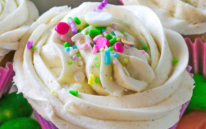 White Chocolate Cupcakes with piped white buttercream frosting, topped with Easter pastel sprinkles in festive pastel Spring colored paper liners, surrounded by brightly colored jelly beans and little fluffy chick and bunny on the side, all sitting on a deep pink cloth © 2026 Jane Bonacci, The Heritage Cook.