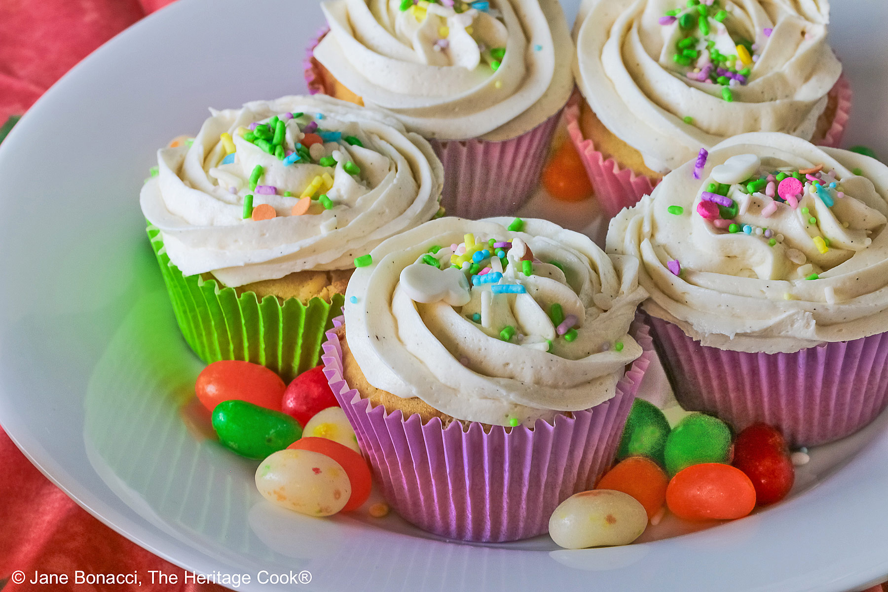 White Chocolate Cupcakes with piped white buttercream frosting, topped with Easter pastel sprinkles in festive pastel Spring colored paper liners, surrounded by brightly colored jelly beans and little fluffy chick and bunny on the side, all sitting on a deep pink cloth © 2026 Jane Bonacci, The Heritage Cook.