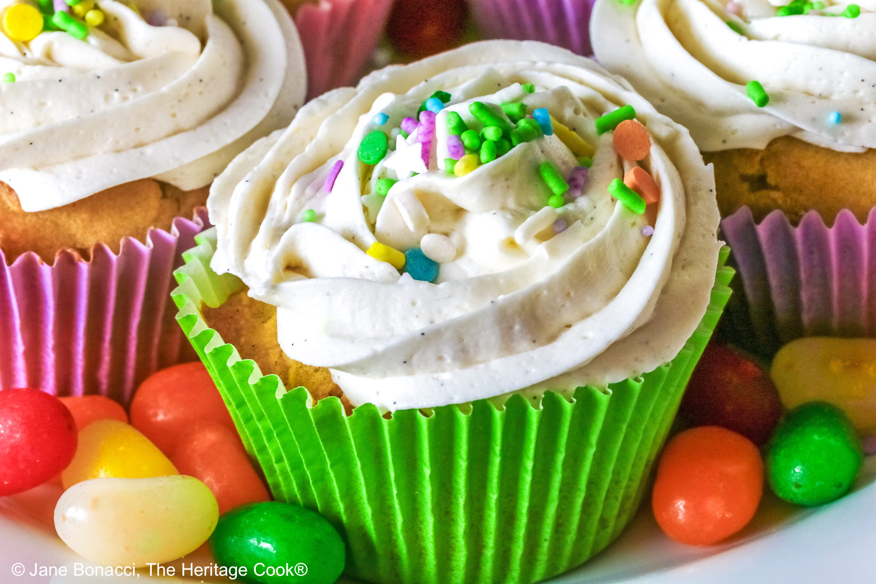 White Chocolate Cupcakes with piped white buttercream frosting, topped with Easter pastel sprinkles in festive pastel Spring colored paper liners, surrounded by brightly colored jelly beans and little fluffy chick and bunny on the side, all sitting on a deep pink cloth © 2026 Jane Bonacci, The Heritage Cook.