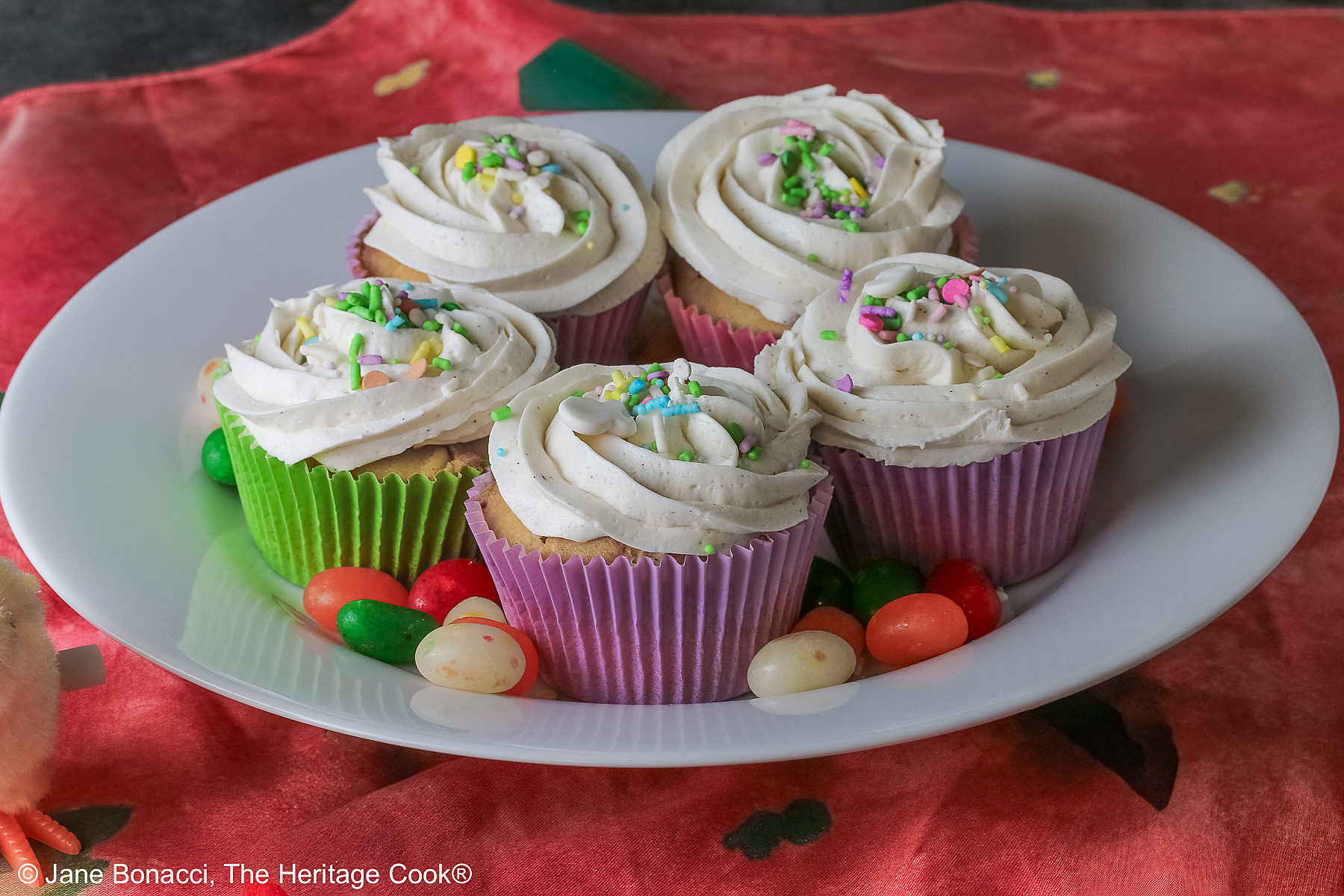 White Chocolate Cupcakes with piped white buttercream frosting, topped with Easter pastel sprinkles in festive pastel Spring colored paper liners, surrounded by brightly colored jelly beans and little fluffy chick and bunny on the side, all sitting on a deep pink cloth © 2026 Jane Bonacci, The Heritage Cook.