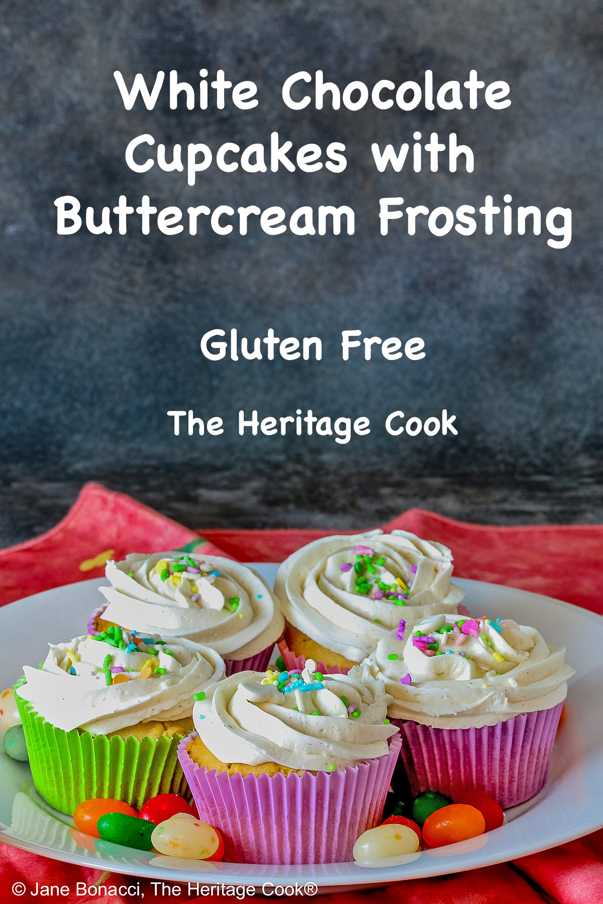 White Chocolate Cupcakes with piped white buttercream frosting, topped with Easter pastel sprinkles in festive pastel Spring colored paper liners, surrounded by brightly colored jelly beans and little fluffy chick and bunny on the side, all sitting on a deep pink cloth © 2026 Jane Bonacci, The Heritage Cook.