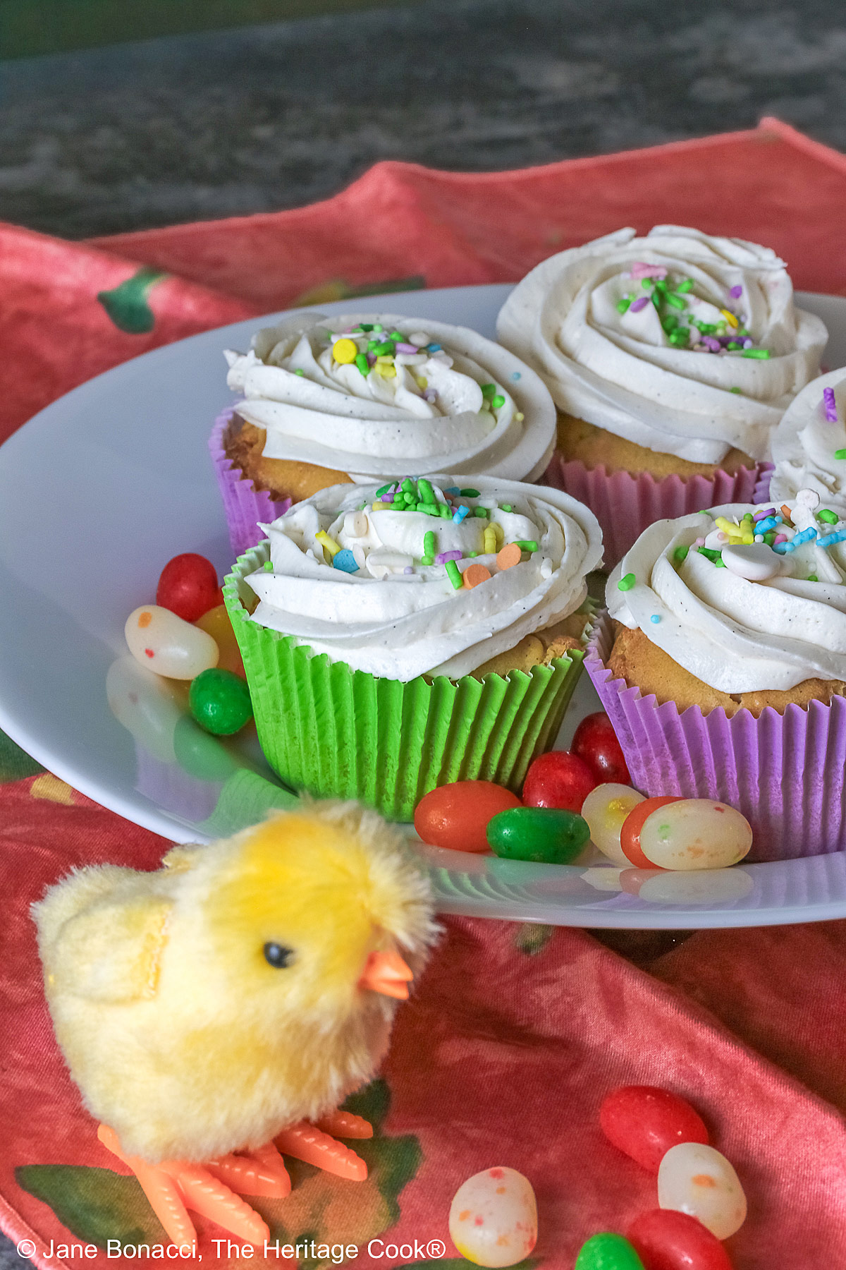 White Chocolate Cupcakes with piped white buttercream frosting, topped with Easter pastel sprinkles in festive pastel Spring colored paper liners, surrounded by brightly colored jelly beans and little fluffy chick and bunny on the side, all sitting on a deep pink cloth © 2026 Jane Bonacci, The Heritage Cook.