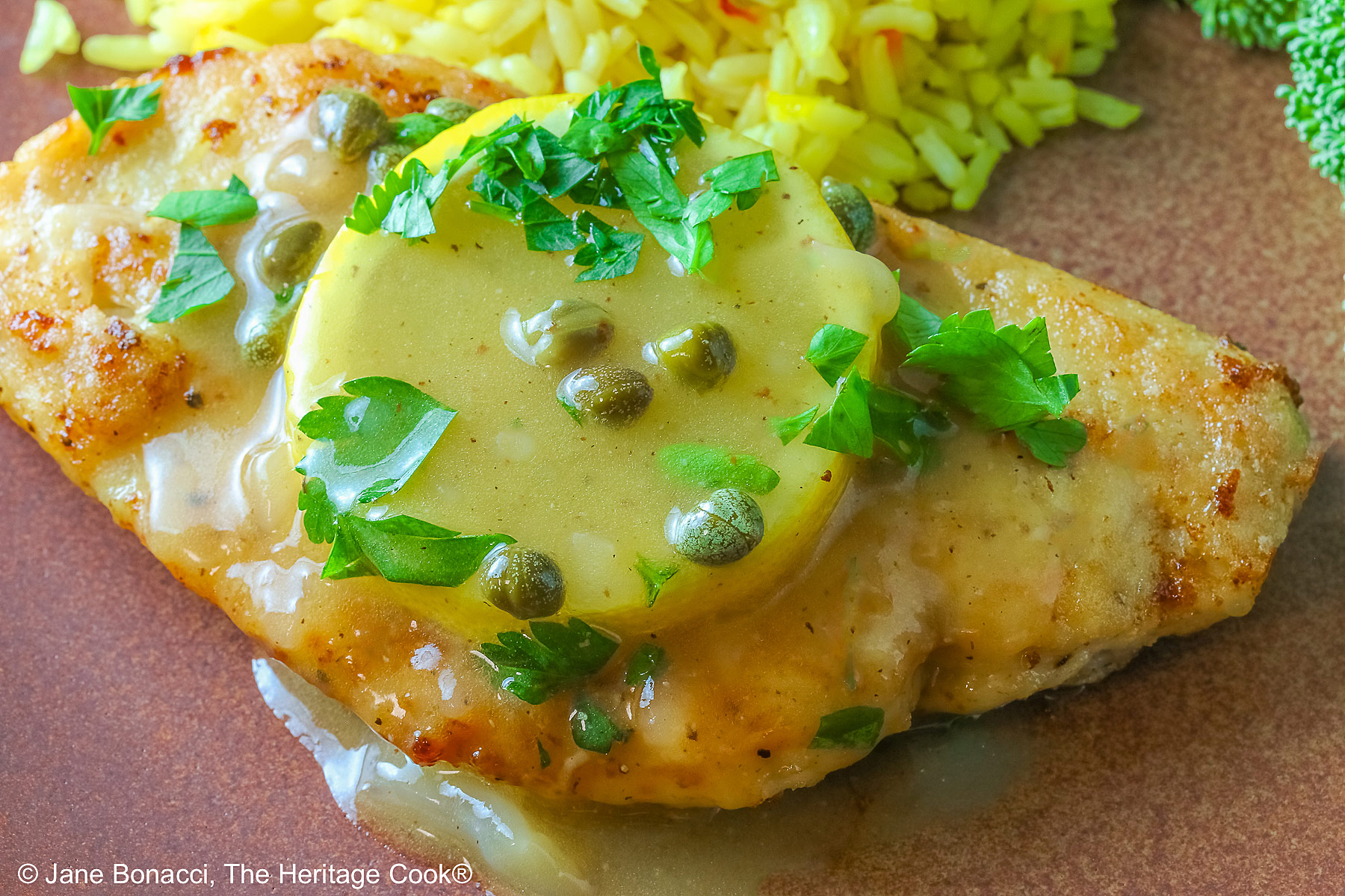 Golden brown chicken cutlet with a lemon slice on top, drizzled with butter-lemon-caper piccata sauce, sprinkled with chopped parsley served with yellow saffron rice and fresh broccoli all on a red’ish brown plate with a blue cloth behind © 2026 Jane Bonacci, The Heritage Cook.