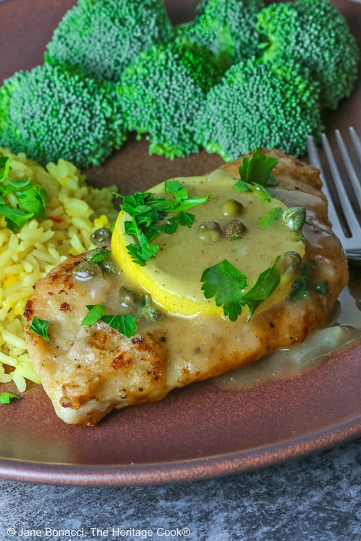 Golden brown chicken cutlet with a lemon slice on top, drizzled with butter-lemon-caper piccata sauce, sprinkled with chopped parsley served with yellow saffron rice and fresh broccoli all on a red’ish brown plate with a blue cloth behind © 2026 Jane Bonacci, The Heritage Cook.