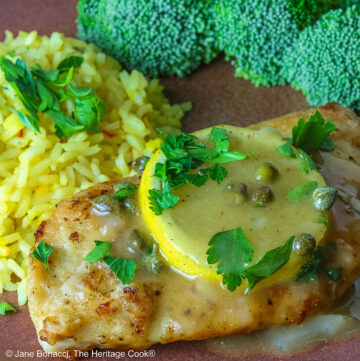 Golden brown chicken cutlet with a lemon slice on top, drizzled with butter-lemon-caper piccata sauce, sprinkled with chopped parsley served with yellow saffron rice and fresh broccoli all on a red’ish brown plate with a blue cloth behind © 2026 Jane Bonacci, The Heritage Cook.