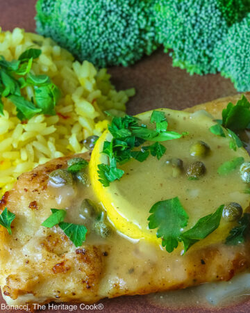 Golden brown chicken cutlet with a lemon slice on top, drizzled with butter-lemon-caper piccata sauce, sprinkled with chopped parsley served with yellow saffron rice and fresh broccoli all on a red’ish brown plate with a blue cloth behind © 2026 Jane Bonacci, The Heritage Cook.