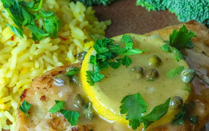 Golden brown chicken cutlet with a lemon slice on top, drizzled with butter-lemon-caper piccata sauce, sprinkled with chopped parsley served with yellow saffron rice and fresh broccoli all on a red’ish brown plate with a blue cloth behind © 2026 Jane Bonacci, The Heritage Cook.