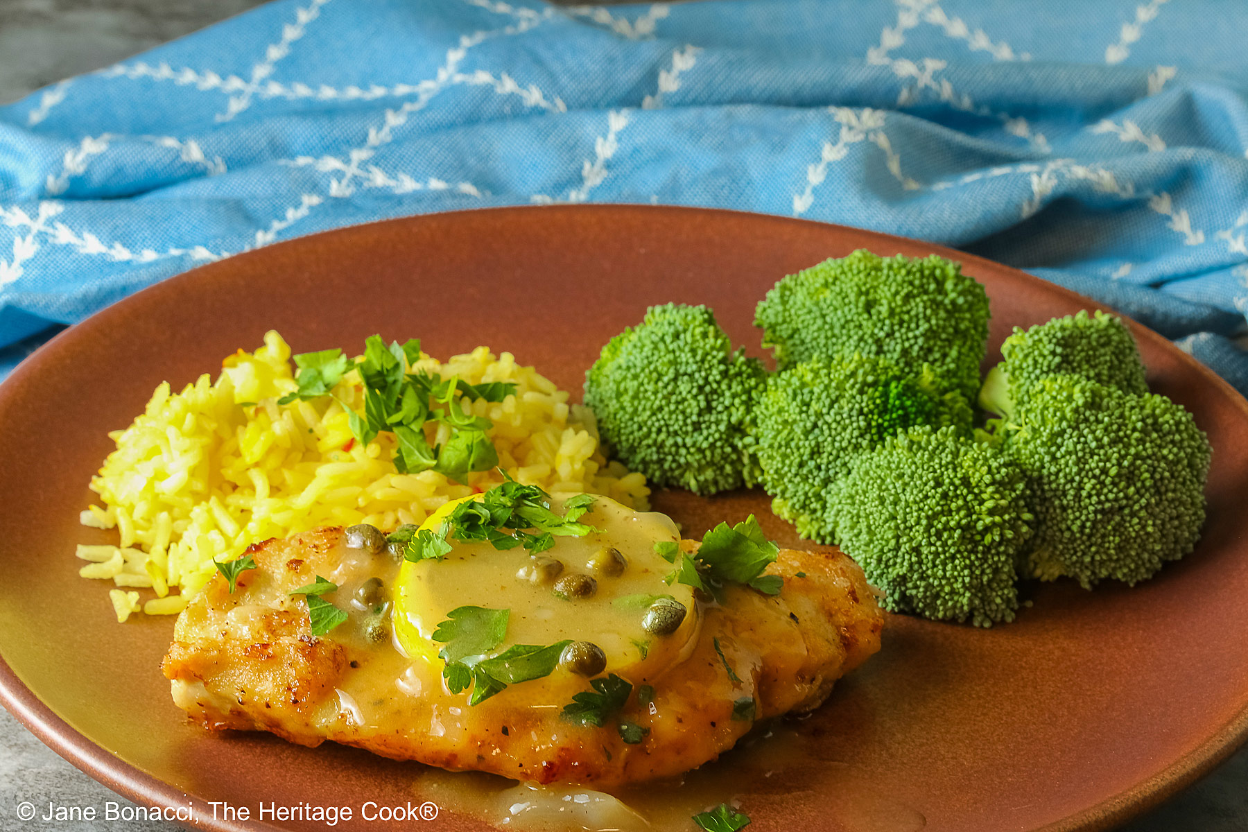 Golden brown chicken cutlet with a lemon slice on top, drizzled with butter-lemon-caper piccata sauce, sprinkled with chopped parsley served with yellow saffron rice and fresh broccoli all on a red’ish brown plate with a blue cloth behind © 2026 Jane Bonacci, The Heritage Cook.