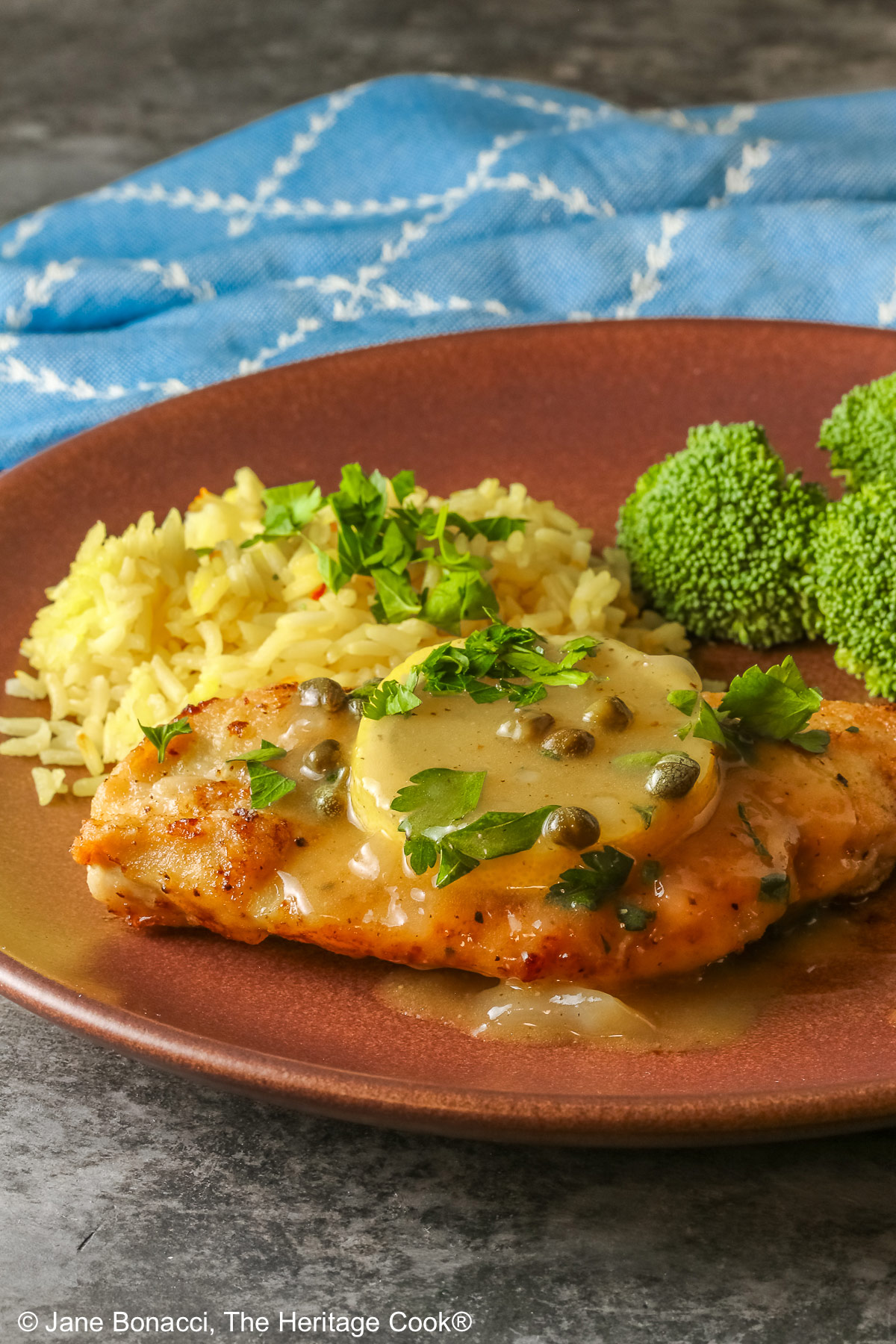 Golden brown chicken cutlet with a lemon slice on top, drizzled with butter-lemon-caper piccata sauce, sprinkled with chopped parsley served with yellow saffron rice and fresh broccoli all on a red’ish brown plate with a blue cloth behind © 2026 Jane Bonacci, The Heritage Cook.