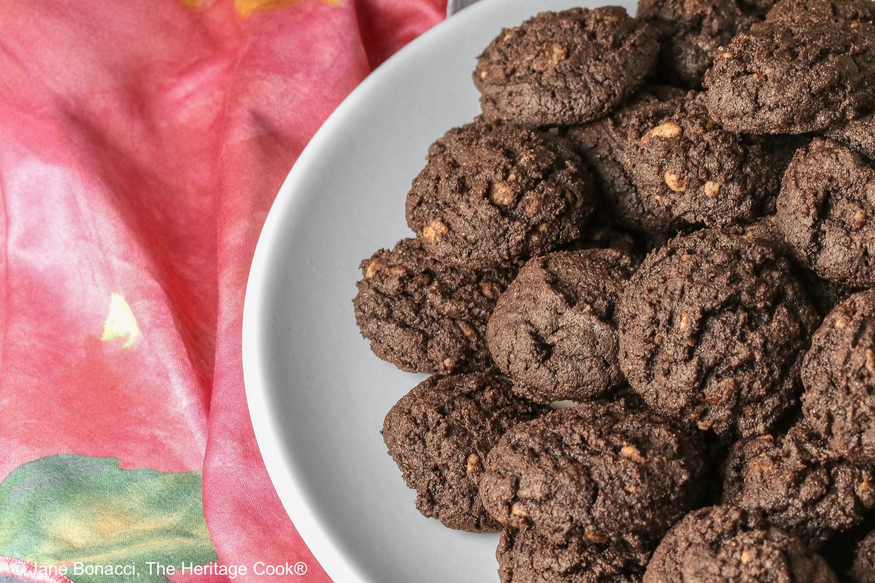 A large pile of very dark crispy chocolate cookies heaped on a cream colored plate, sitting on a pink cloth; some with a small white plate in front with 3 individual cookies on it © 2026 Jane Bonacci, The Heritage Cook.