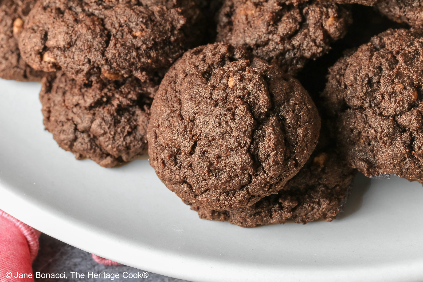 A large pile of very dark crispy chocolate cookies heaped on a cream colored plate, sitting on a pink cloth; some with a small white plate in front with 3 individual cookies on it © 2026 Jane Bonacci, The Heritage Cook.