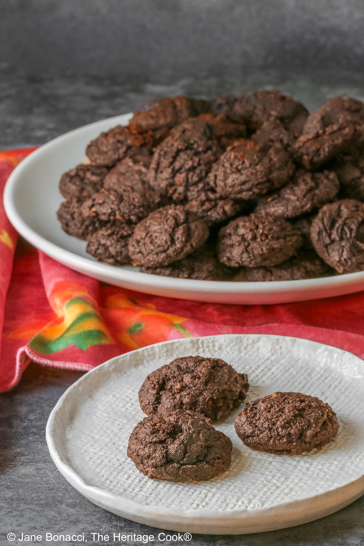 A large pile of very dark crispy chocolate cookies heaped on a cream colored plate, sitting on a pink cloth; some with a small white plate in front with 3 individual cookies on it © 2026 Jane Bonacci, The Heritage Cook.