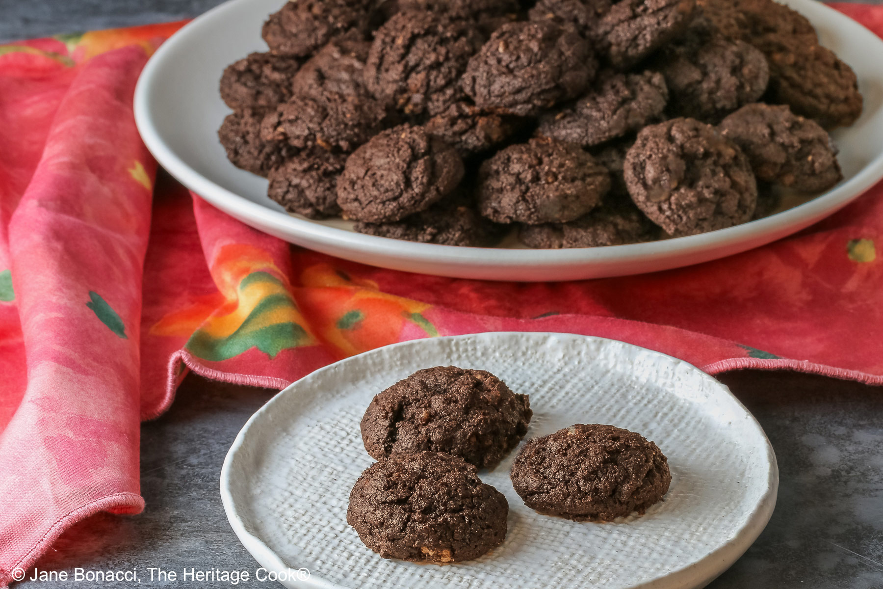 A large pile of very dark crispy chocolate cookies heaped on a cream colored plate, sitting on a pink cloth; some with a small white plate in front with 3 individual cookies on it © 2026 Jane Bonacci, The Heritage Cook.