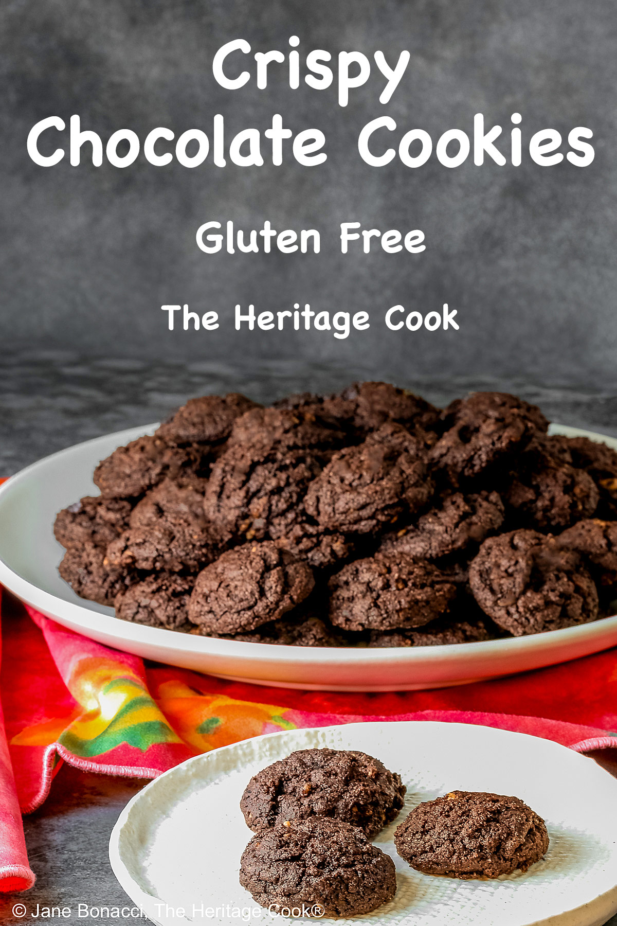 A large pile of very dark crispy chocolate cookies heaped on a cream colored plate, sitting on a pink cloth; some with a small white plate in front with 3 individual cookies on it © 2026 Jane Bonacci, The Heritage Cook.