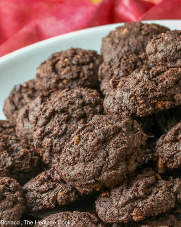 A large pile of very dark crispy chocolate cookies heaped on a cream colored plate, sitting on a pink cloth; some with a small white plate in front with 3 individual cookies on it © 2026 Jane Bonacci, The Heritage Cook.