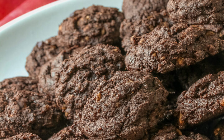 A large pile of very dark crispy chocolate cookies heaped on a cream colored plate, sitting on a pink cloth; some with a small white plate in front with 3 individual cookies on it © 2026 Jane Bonacci, The Heritage Cook.