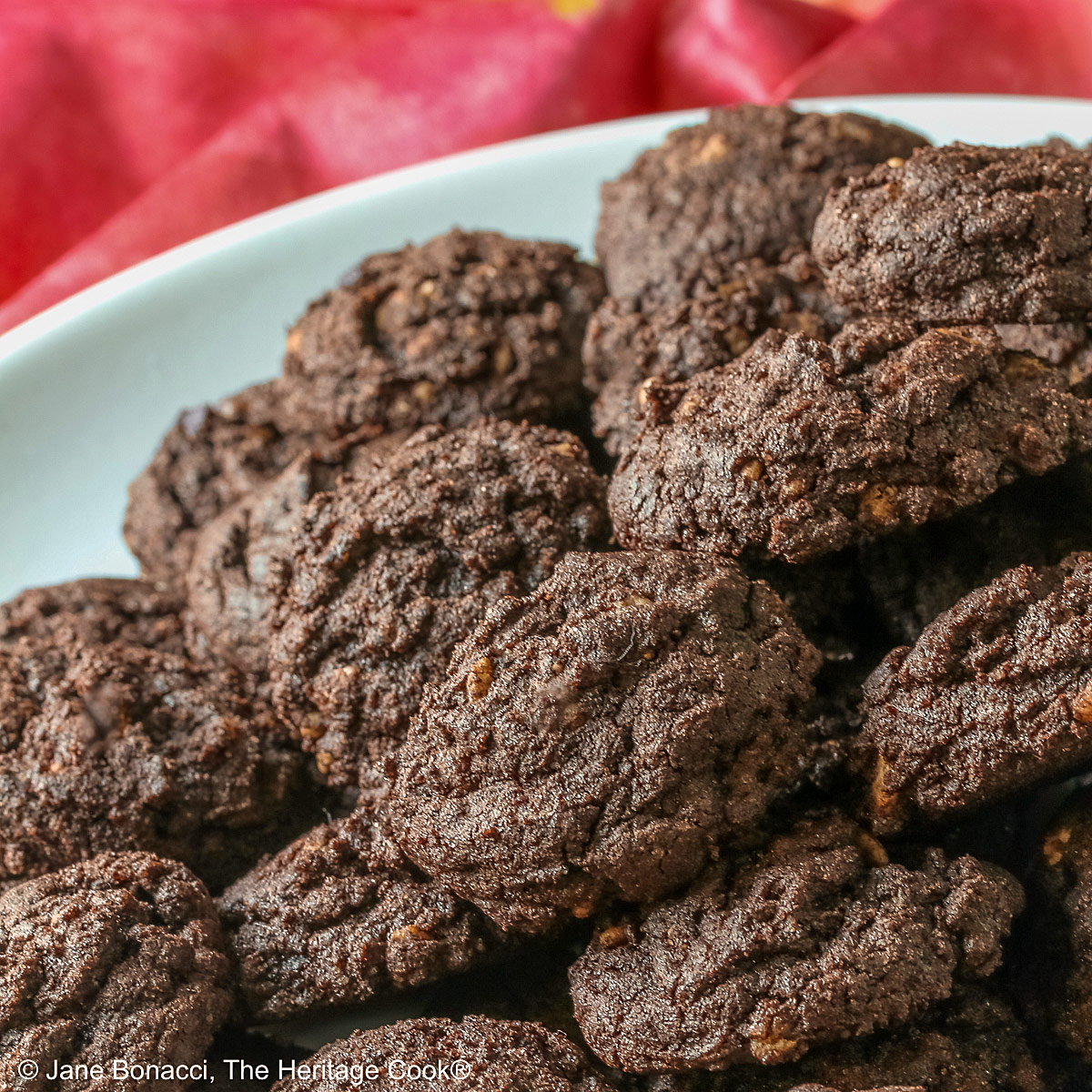 A large pile of very dark crispy chocolate cookies heaped on a cream colored plate, sitting on a pink cloth; some with a small white plate in front with 3 individual cookies on it © 2026 Jane Bonacci, The Heritage Cook.