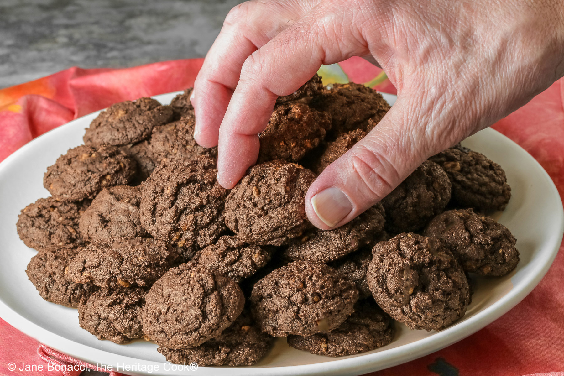 A man's hand reaching for a cookie on the top of the pile of chocolate cookies.