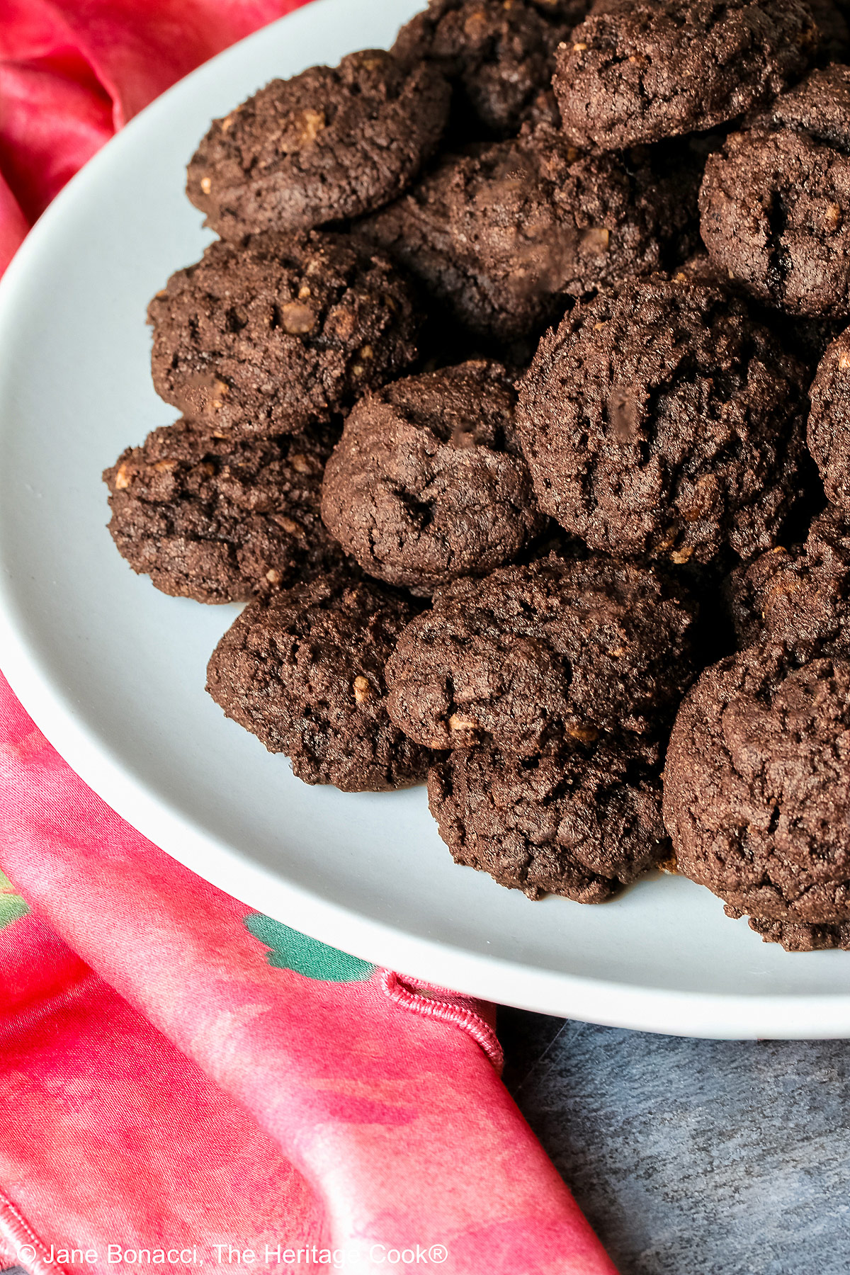 A large pile of very dark crispy chocolate cookies heaped on a cream colored plate, sitting on a pink cloth; some with a small white plate in front with 3 individual cookies on it © 2026 Jane Bonacci, The Heritage Cook.
