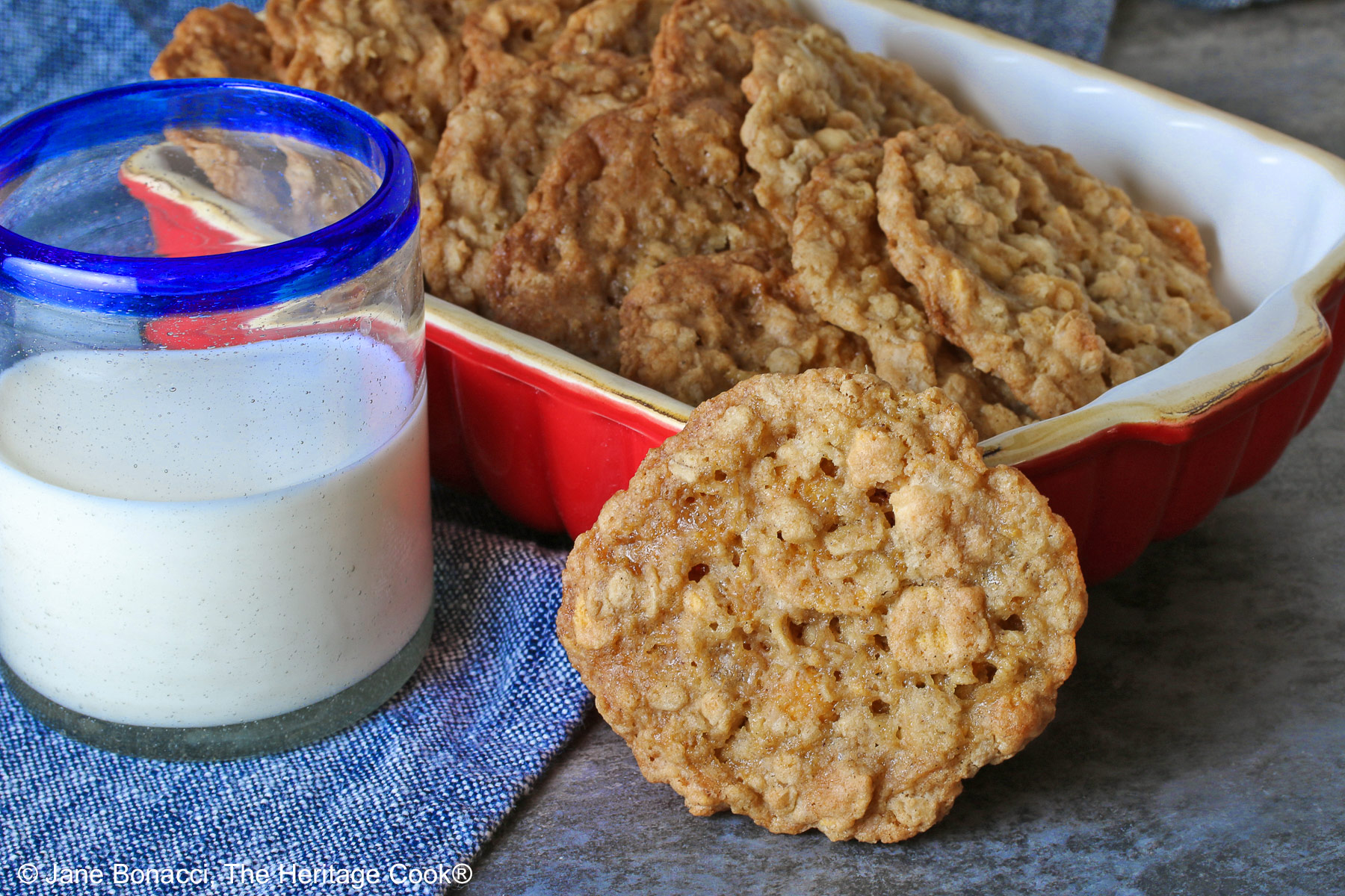 Golden brown Crunchy Oatmeal Cookies are standing upright in a small red baking dish, leaning against each other, with a glass of milk beside it and a single cookie propped up against the front of the dish. All sitting on a blue cloth with a gray background behind © 2026 Jane Bonacci, The Heritage Cook.