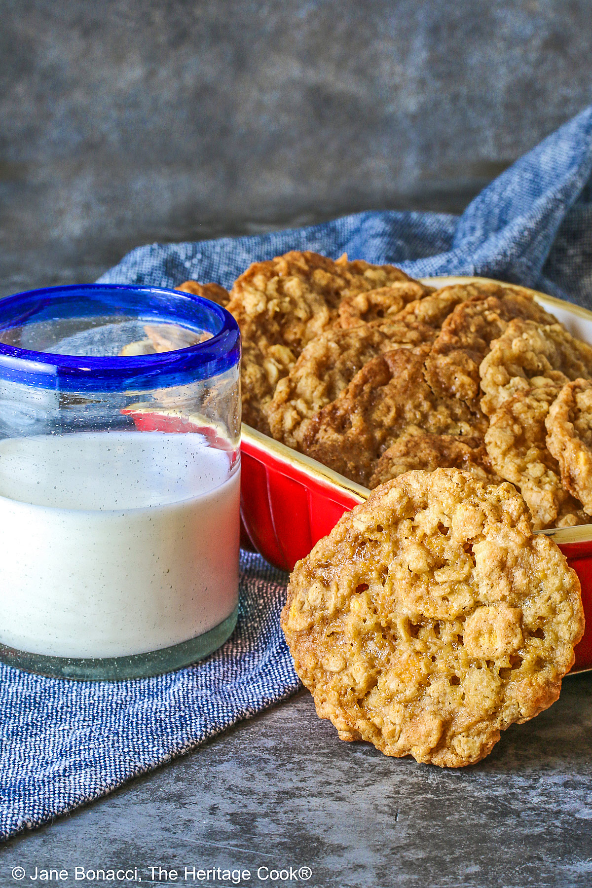 Golden brown Crunchy Oatmeal Cookies are standing upright in a small red baking dish, leaning against each other, with a glass of milk beside it and a single cookie propped up against the front of the dish. All sitting on a blue cloth with a gray background behind © 2026 Jane Bonacci, The Heritage Cook.