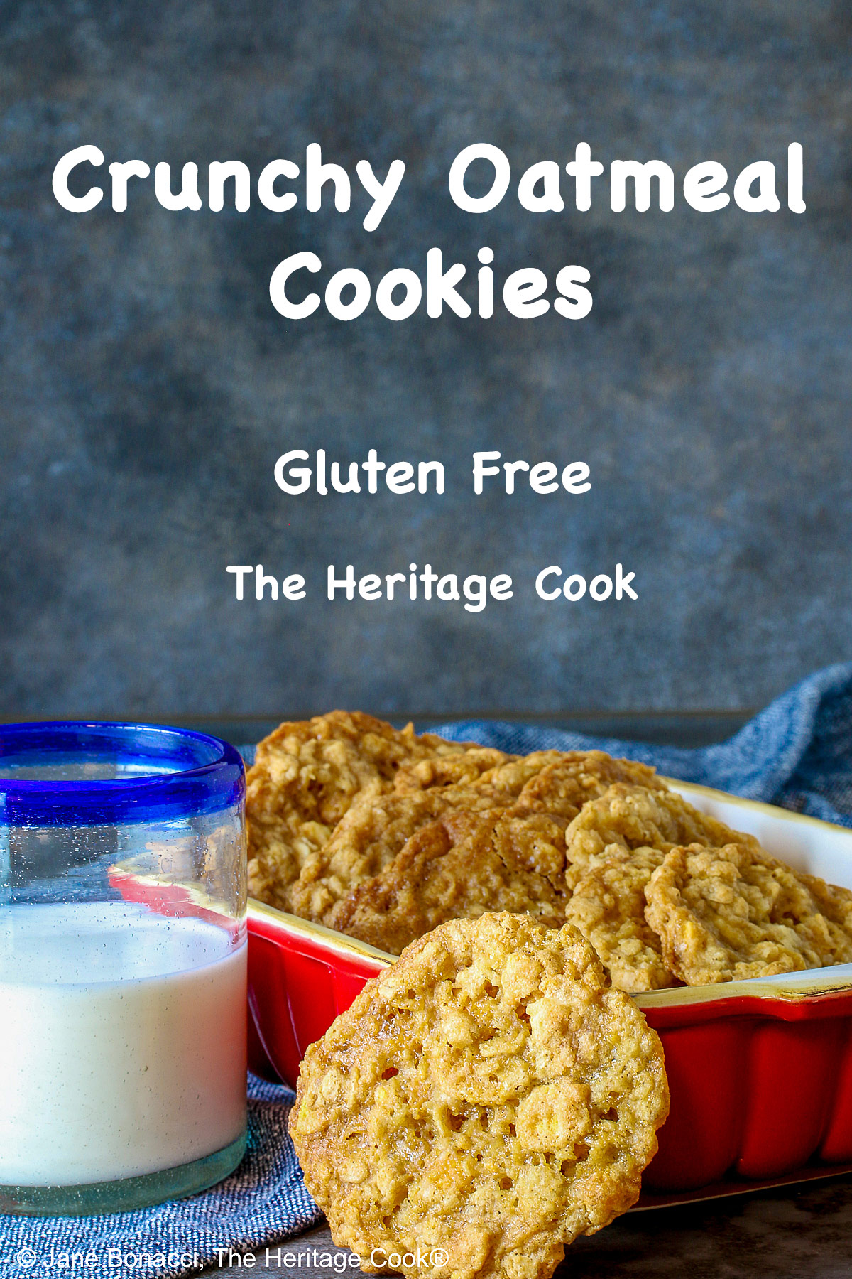 Golden brown Crunchy Oatmeal Cookies are standing upright in a small red baking dish, leaning against each other, with a glass of milk beside it and a single cookie propped up against the front of the dish. All sitting on a blue cloth with a gray background behind © 2026 Jane Bonacci, The Heritage Cook.