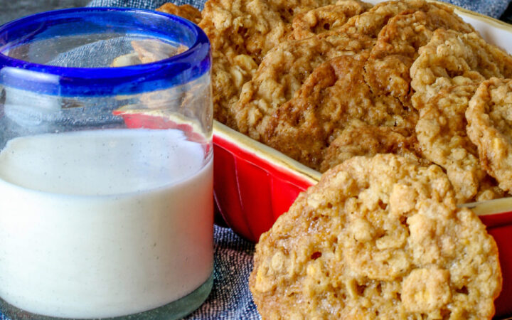 Golden brown Crunchy Oatmeal Cookies are standing upright in a small red baking dish, leaning against each other, with a glass of milk beside it and a single cookie propped up against the front of the dish. All sitting on a blue cloth with a gray background behind © 2026 Jane Bonacci, The Heritage Cook.